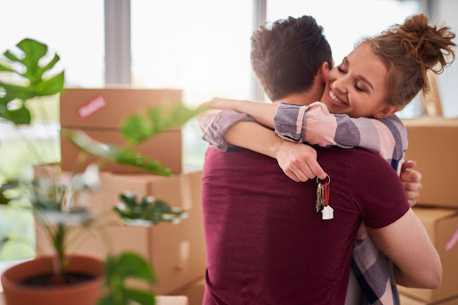 mulher abraçando companheiro com uma chave com pingente de casa em mãos e sorrindo. Ao fundo caixa de mudanças e um vaso com plantas